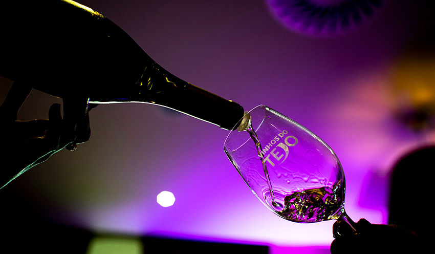 Mão serve vinho em taça contra luz roxa. A taça tem a inscrição Vinhos do Tejo. Ambiente elegante e descontraído.