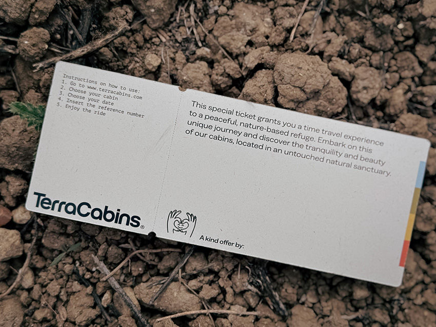 Ingresso branco no solo seco, com instruções e logotipo "TerraCabins". Texto menciona uma experiência de viagem na natureza.