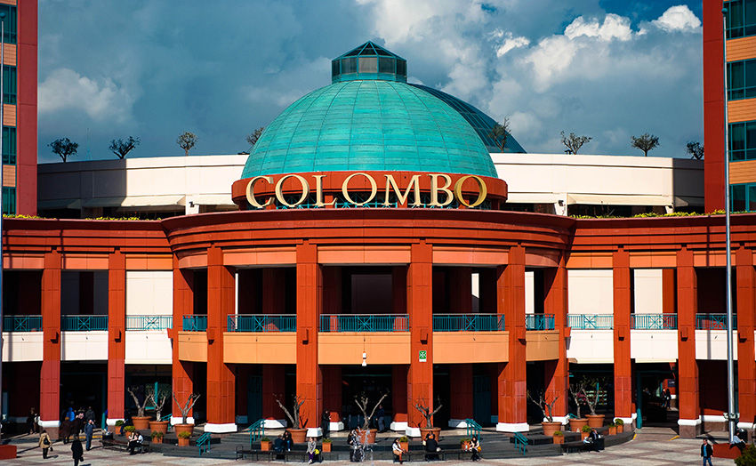 Shopping Colombo com cúpula azul e fachada terracota. Pessoas caminham na entrada. Céu azul com nuvens ao fundo.