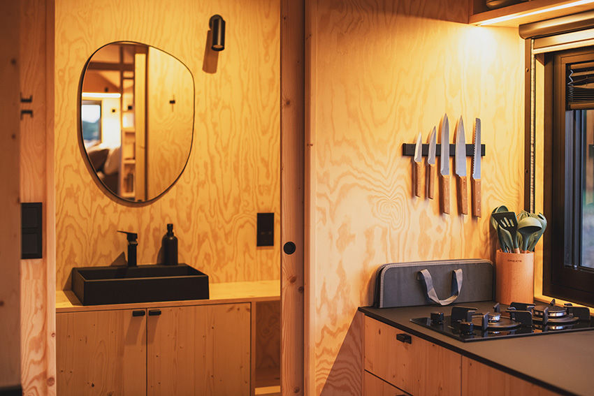 Interior de cozinha minimalista em madeira clara. Conjunto de facas na parede, pia preta e espelho, criando atmosfera aconchegante.