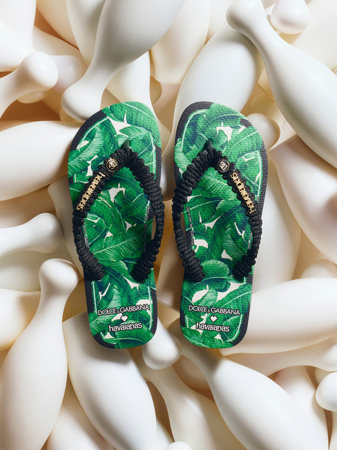 Chinelos verdes com estampa de folhas e tiras pretas sobre fundo de formas brancas. Logo "Dolce & Gabbana" e "Havaianas" visível.