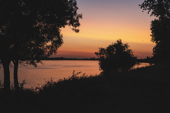 Pôr do sol sobre rio tranquilo, com árvores escuras em silhueta e céu alaranjado. Atmosfera serena e natural.