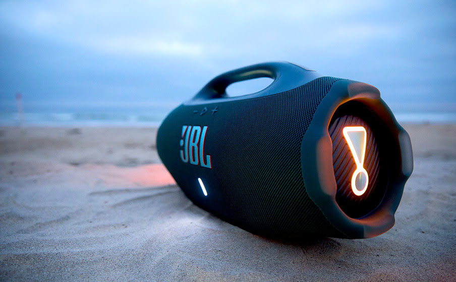 Caixa de som JBL preta na areia da praia ao anoitecer. Luz acesa em amarelo na lateral, céu azul acinzentado ao fundo.