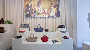 Exposição de bolsas em várias cores sobre prateleiras brancas. Ao fundo, uma foto de pessoas em uma festa, com cortinas atrás.