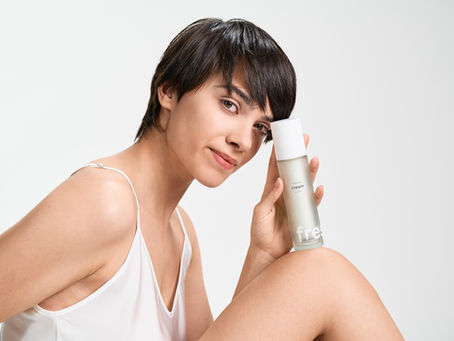 Mulher de cabelo curto sorri enquanto segura um frasco transparente de creme "fresh" em fundo branco. Veste roupa branca, transmitindo leveza.