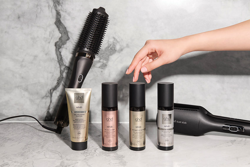 Mão pegando frasco de produto "ghd rehab" em bancada de mármore. Ao lado, escova elétrica e outros produtos de cabelo. Fundo cinza claro.