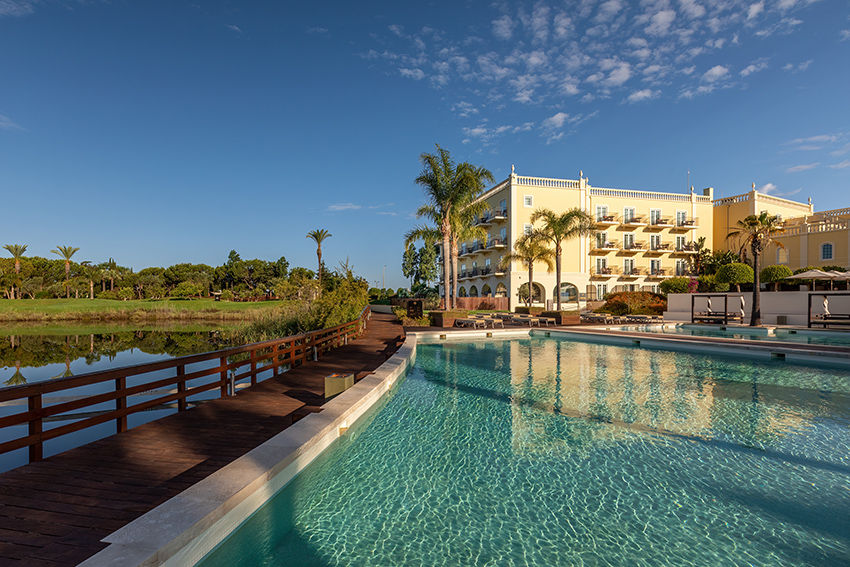 Edifício amarelo com palmeiras ao lado de piscina azul clara. Céu limpo, ponte de madeira, natureza verde ao fundo, atmosfera tranquila.