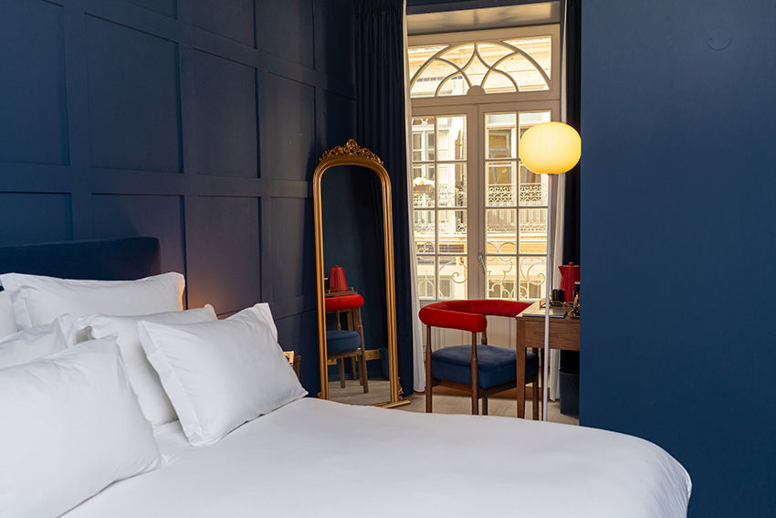 Quarto com paredes azuis, espelho de moldura dourada, cama branca, cadeira vermelha e luminária amarela. Ambiente elegante e acolhedor.