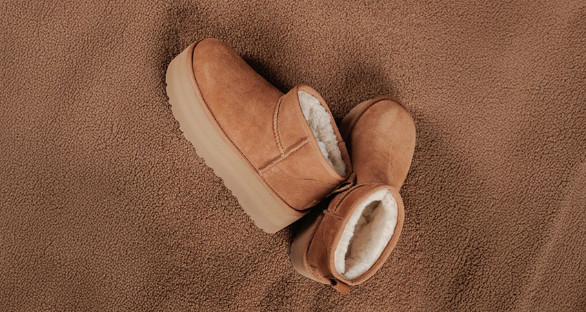 Botas de camurça marrom claro com forro branco estão sobre uma superfície marrom texturizada, criando um tom monocromático acolhedor.