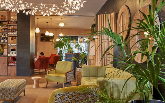 Sala de estar moderna com sofá verde, poltronas estampadas, plantas, luminárias pendentes e estantes de bebidas ao fundo. Ambiente acolhedor.
