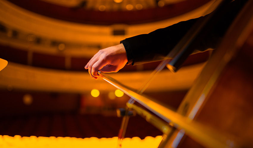 Mão tocando violoncelo em teatro iluminado com luzes quentes. Fundo desfocado com assentos vazios. Atmosfera enfática e artística.