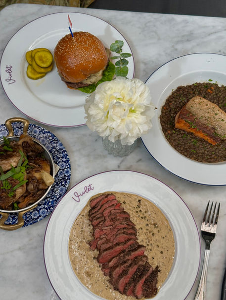 A marble table features a variety of dishes from Violet Bistro including the Smash Burger, Steak au Povre, Salmon on Lentils, and Roasted Veg