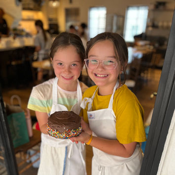 Two little girls hold a chocolate cake they made at Violet Cooking School