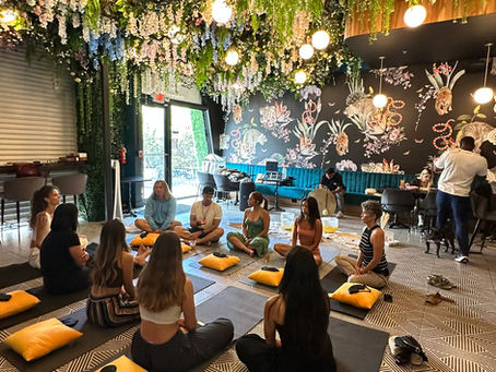People sit in a circle on yoga mats with cushions, in a lush, plant-decorated room with patterned floors and hanging lights, creating a serene mood.