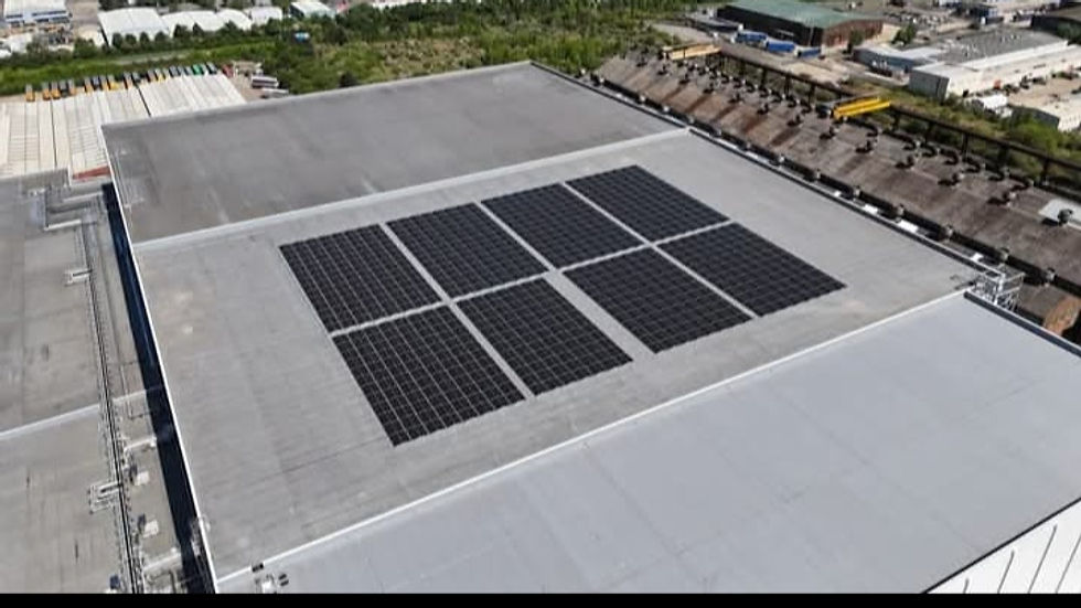 Aerial view of a large warehouse roof with black solar panels in a grid pattern. Green industrial area surrounds the building.