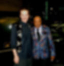 Two men smiling at night in front of a car. One wears a patterned vest, the other a floral blazer. Sign reads "POWER & LIGHT."