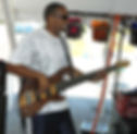 Man playing bass guitar under a tent, smiling and wearing sunglasses. Colorful lights hang above, bright day in the background, relaxed mood.