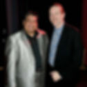 Two men smiling, one in a shiny silver suit, the other in a black jacket with a blue patterned shirt. Dimly lit indoor setting.