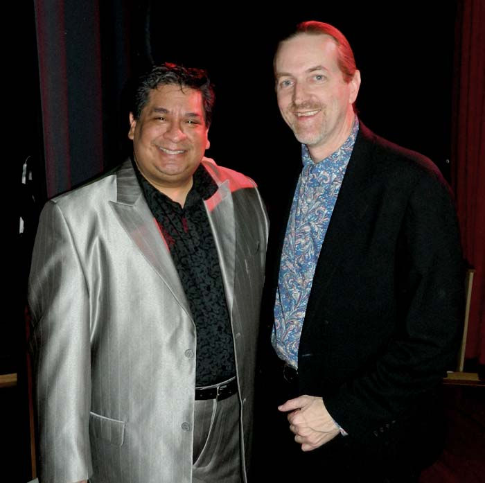 Two men smiling, one in a shiny silver suit, the other in a black jacket with a blue patterned shirt. Dimly lit indoor setting.