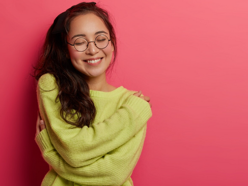 หญิงสาวสวมแว่นยิ้มหลับตาและโอบกอดตัวเองบนพื้นหลังสีชมพู สื่อถึงความรู้สึกปลอดภัยทางอารมณ์ (Emotional Safety) และการเคารพสิทธิ์ในเนื้อตัวร่างกายตัวเอง (Agency)