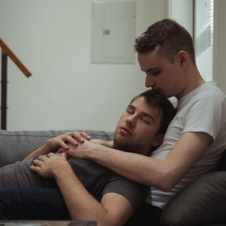 คู่รักชายรักชาย (Gay Couple) นอนโอบกอดและกุมมือกันบนโซฟาอย่างผ่อนคลาย สื่อถึงความรู้สึกมั่นคง การทำความเข้าใจอารมณ์ความรู้สึก และการดูแลโลกภายในใจของคนรัก