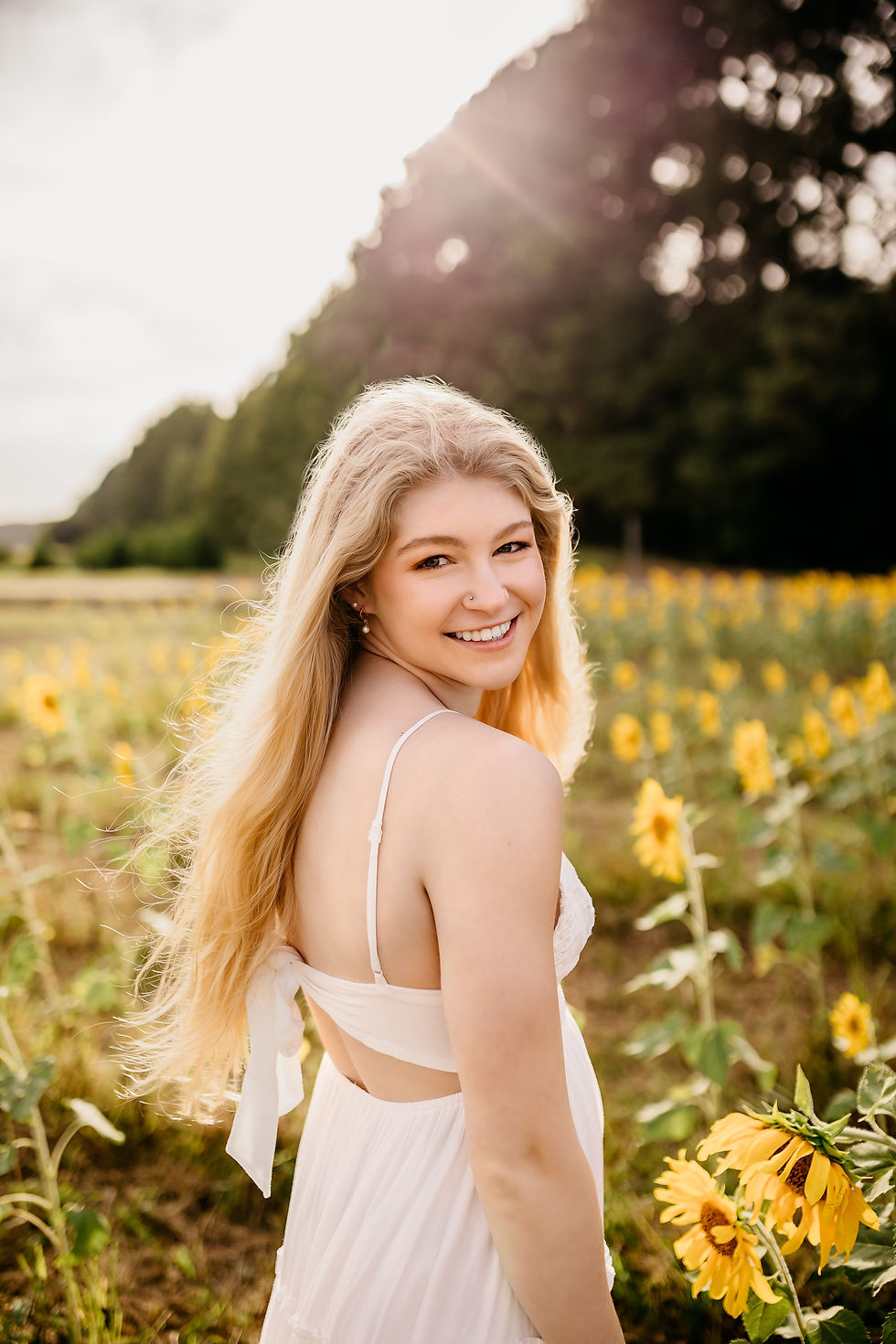 Smiling woman in a white dress stands in a sunflower field, under bright sunlight. Lush green trees form the background, creating a serene mood.