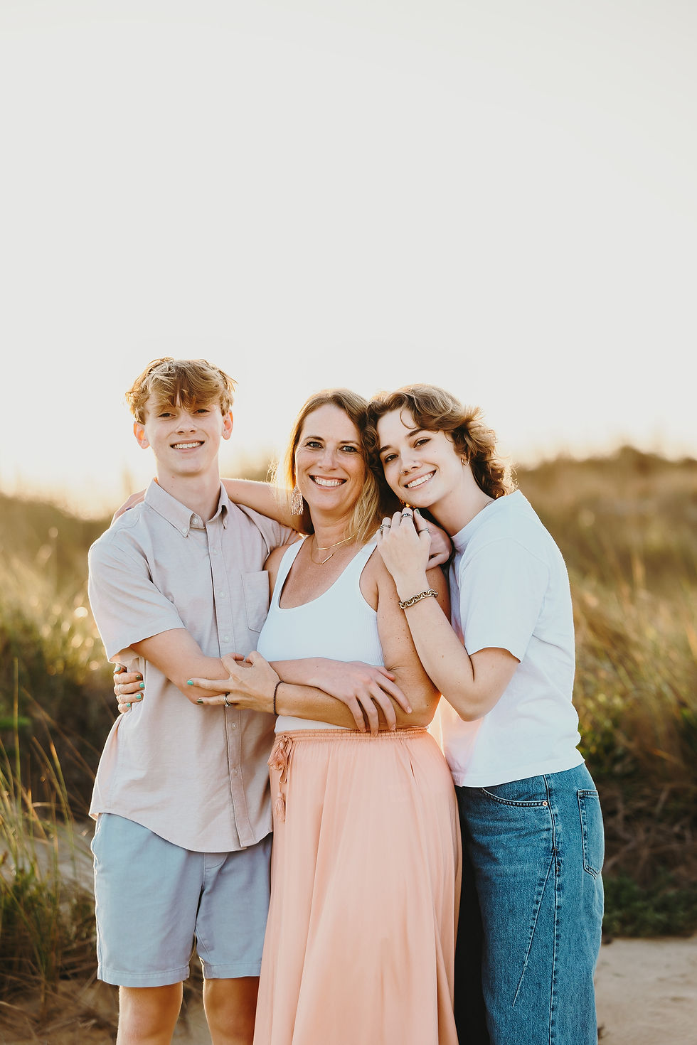 Smiling woman embraces two teens in casual outfits, standing in a sunlit grassy field. The mood is joyful and warm.