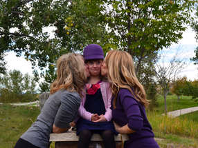 Two woman in purple kissing the cheeks of a little girl in a purple hat outdoors.