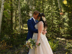 Groom and bride embracing each other  face to face in front of tall green trees.
