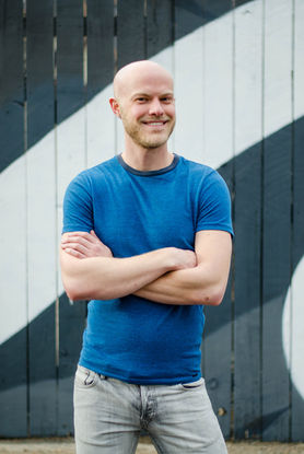 Man in blue tshirt and jeans posing with arms crossed and smiling while standing in front of painted mural fence.