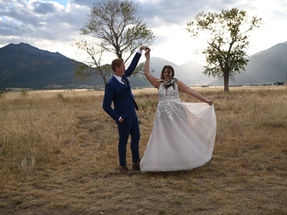 Groom and bride dancing in open dry field with single trees in the background and mountains with the sun setting.