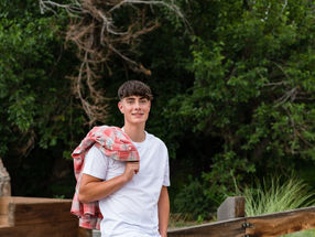 Young man in a white tshirt and jeans with red flannel thrown over his shoulder and leaning on fence post near the woods.