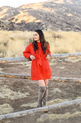 Young woman in red cropped long sleeve dressed leaned against a wood fence near the mountains.