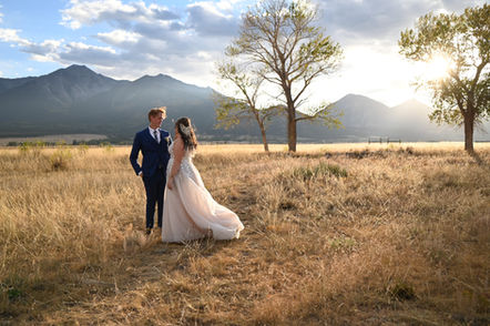 Groom and bride standing in open dry field with single trees in the background and mountains with the sun setting.