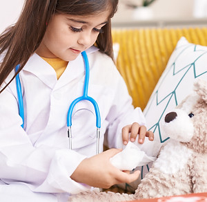 Adorable hispanic girl wearing doctor uniform bandaging teddy bear arm at home.jpg