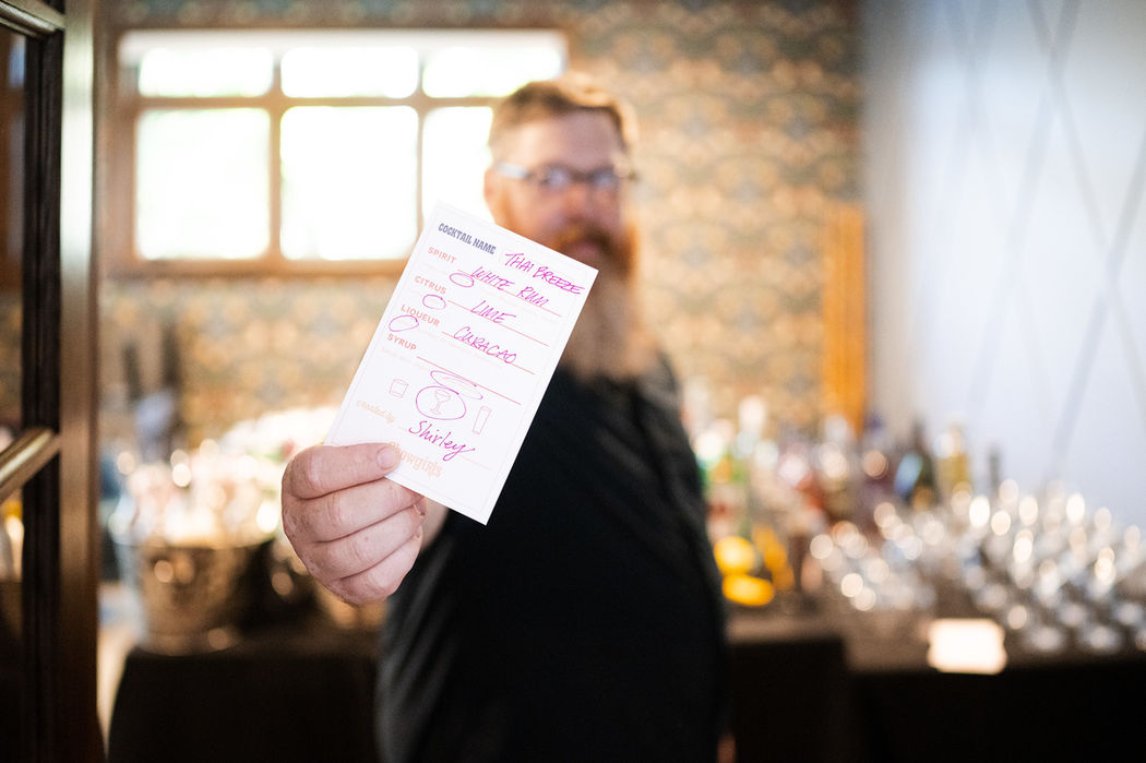 A Chowgirls bartender hold a custom drink card up to the camera. There is a bartender workstation set up in the background. The inmage is taken in an urban warehouse style venue.