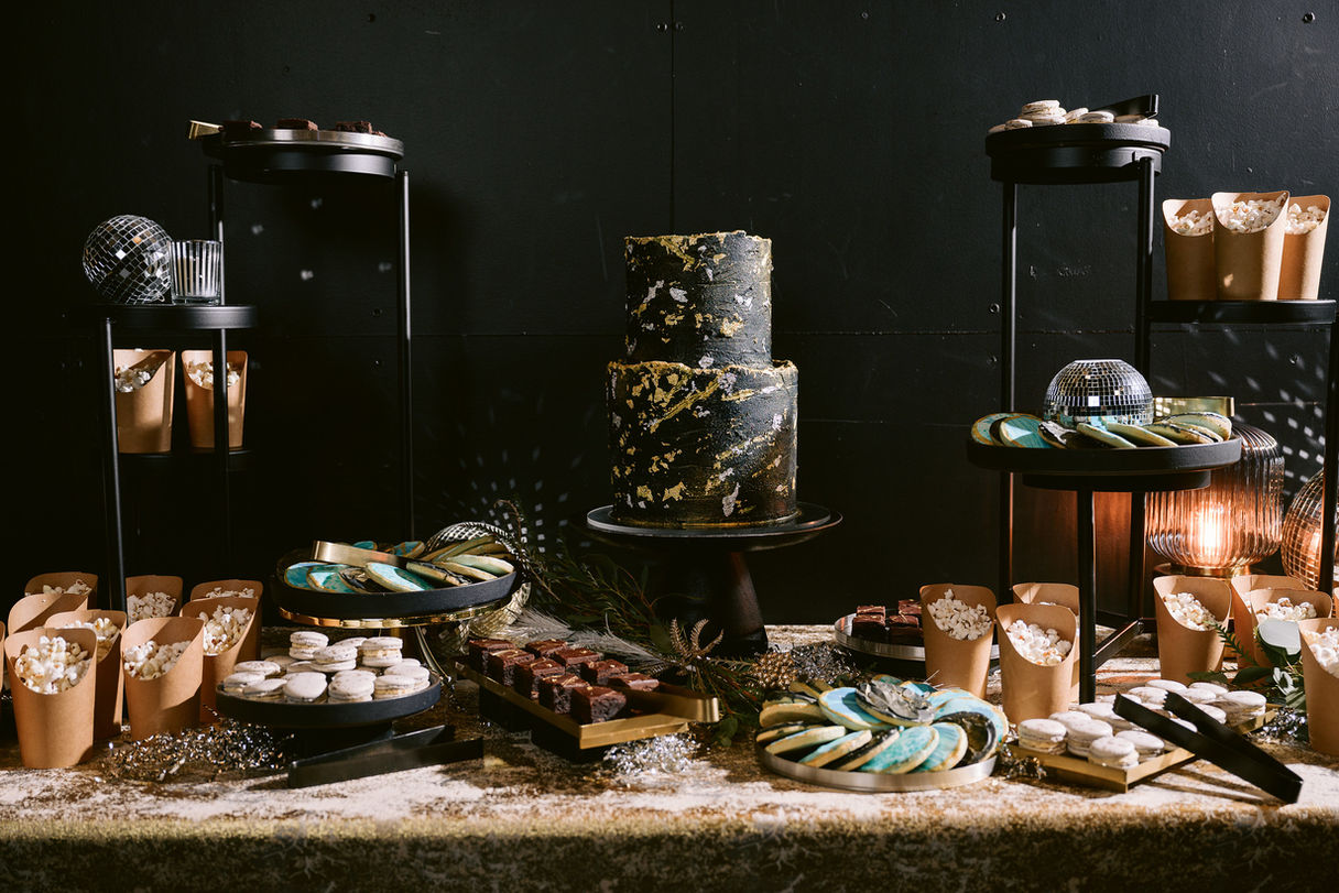 Custom dessert and snack spread designed for a disco-themed event, featuring a dark, shiny color palette with cake, cookies, macarons, and bite-sized brownies.