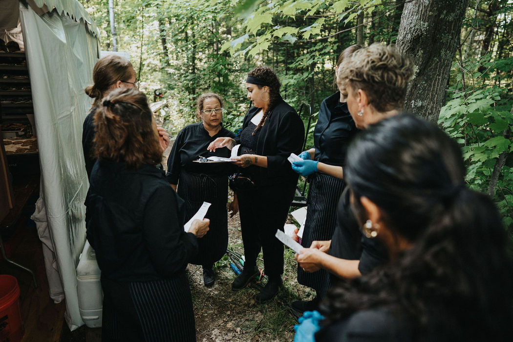 A group of Chowgirls hospitality team members having a pre-shift meeting outside a tent at an outdoor event.