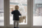 Young toddler with blonde hair and black sweater looks outside of sliding glass doors at a snowy yard.