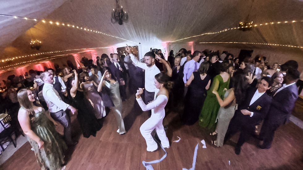 A packed wedding reception dance floor with the bride and groom dancing surrounded by guests in a tent venue with string lights and pink uplighting.