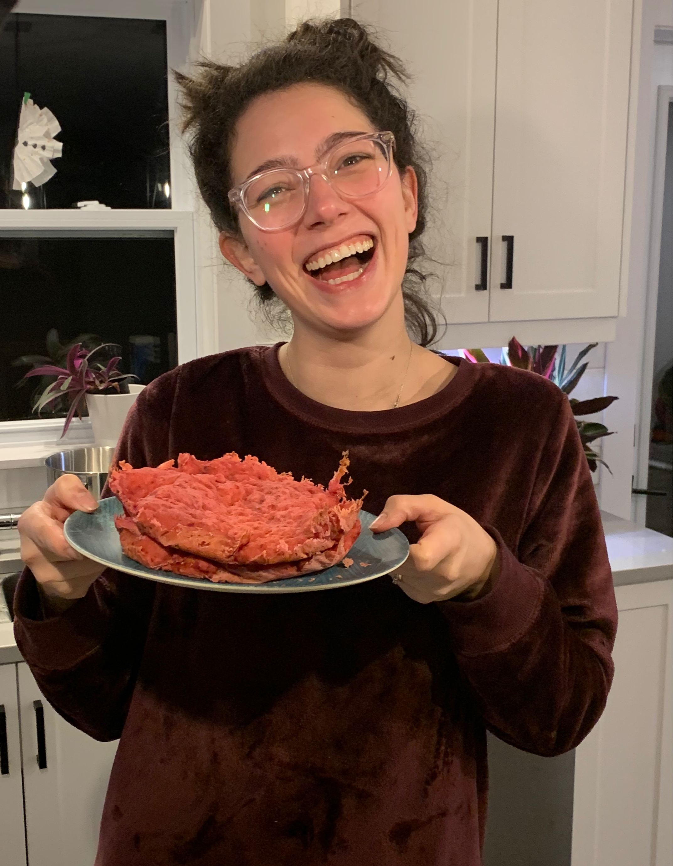 a women smiles while holding a blue plate. on the blue plate is two round slabs of pink cake that look like ground beef.