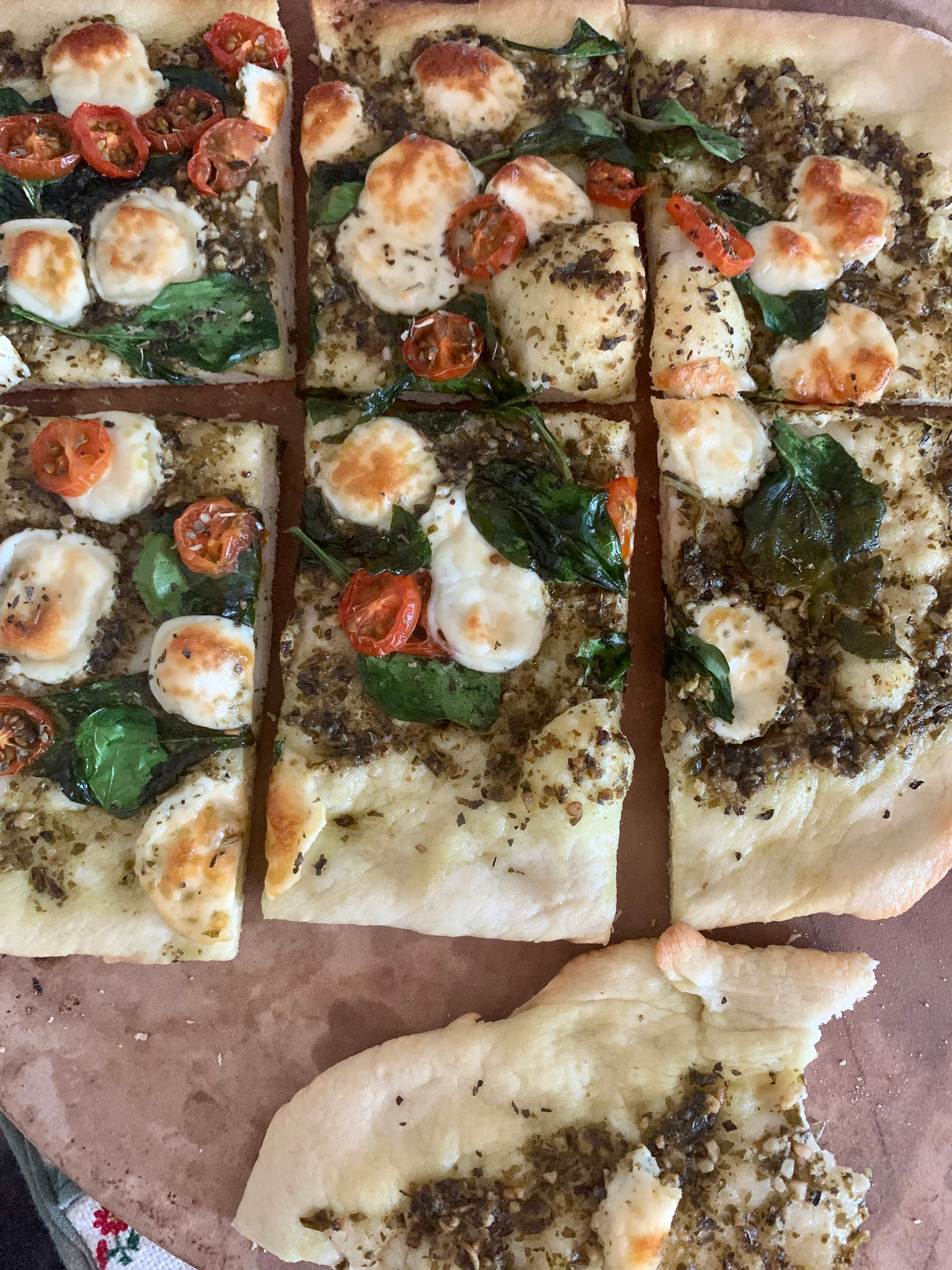 thin crust pizza sits on a pizza stone. there are tomatoes, and spinach, and pesto on the pizza.