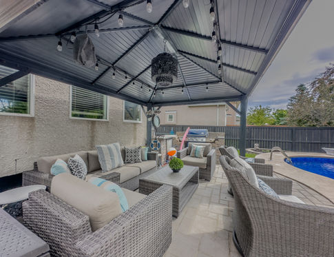 An outdoor patio setup featuring a covered seating area with cushioned wicker furniture, including sofas and chairs arranged around a central coffee table. The space is sheltered by a metal pergola adorned with string lights, creating a cozy atmosphere. The patio is adjacent to a swimming pool, visible in the background, and the overall setting is designed for outdoor relaxation and entertaining.