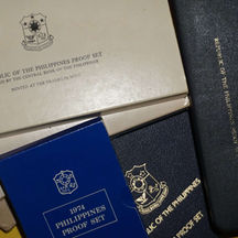 Close-up of multiple Republic of the Philippines proof set cases and folders from the 1970s–1980s, including a blue 1974 Philippines Proof Set box and official Central Bank of the Philippines packaging with embossed national seals and gold lettering.