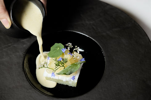 A cream colored soup is poured into a black bowl, where a square piece of squash sits topped with spherical yellow pear, green nasturtium leaf, and small purple flower petals.