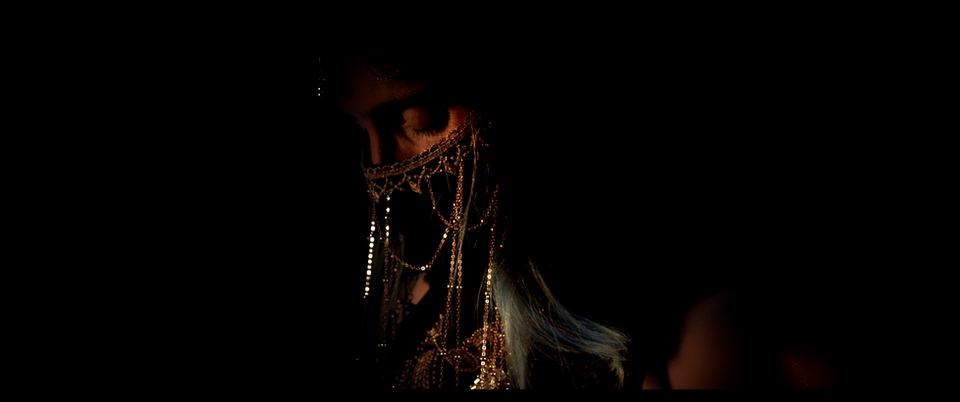 Veiled model wearing layered gold jewellery against a deep black background.