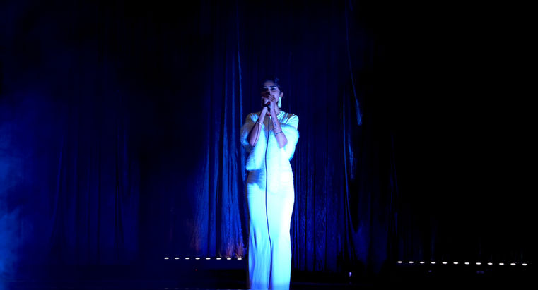 Singer in white performing alone beneath a narrow blue spotlight.