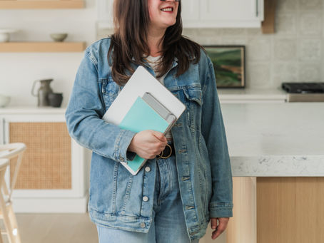 Woman standing in a bright, modern kitchen space holding a tablet and notebook, wearing a denim jacket and jeans, smiling and looking off to the side.