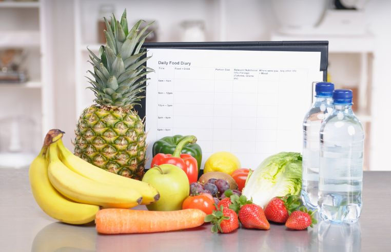 Fresh fruit, vegetables, and water bottles in front of a daily food diary template, representing how keeping a food log can help identify acid reflux triggers.