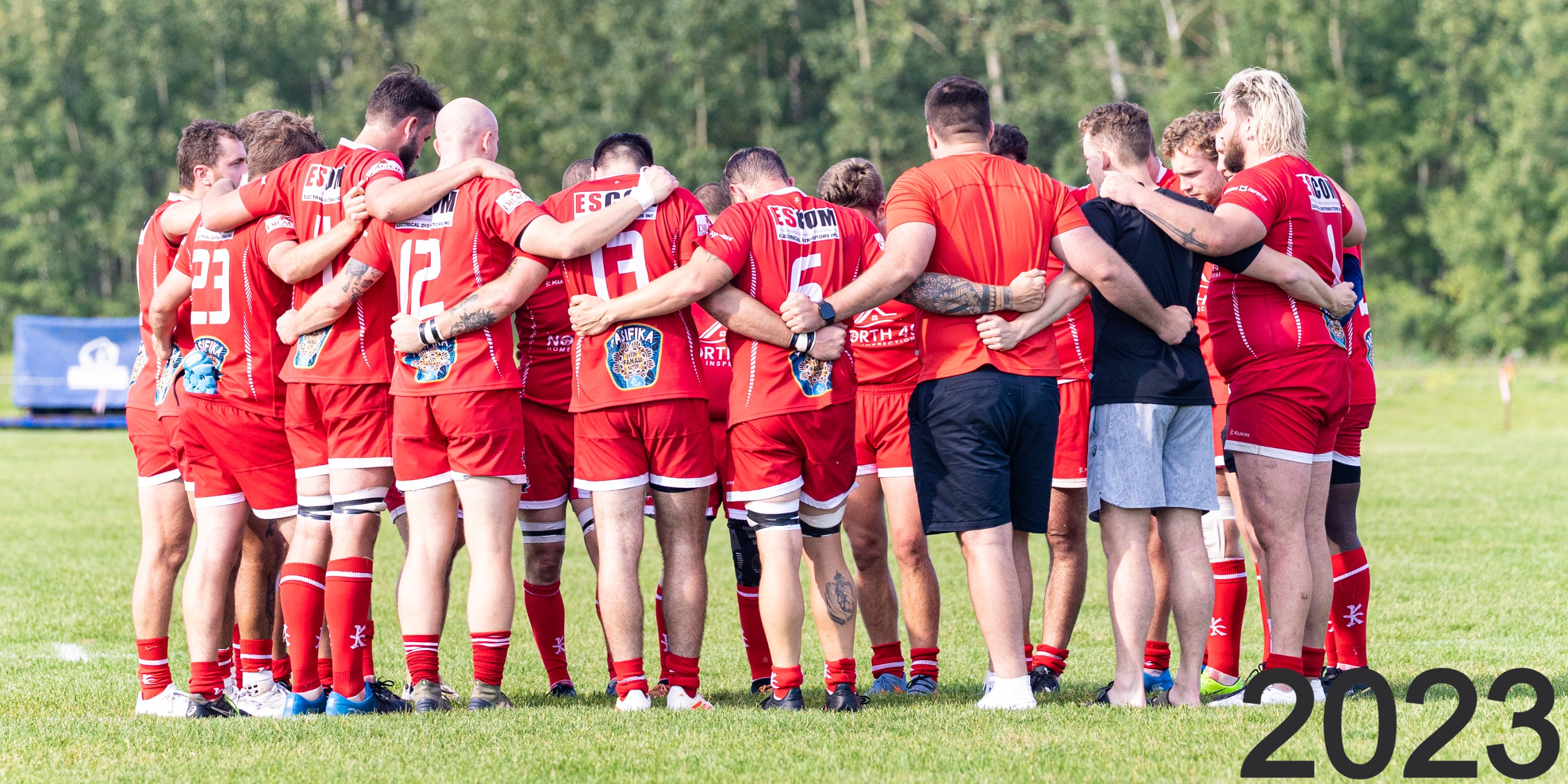 Strathcona Druids senior men's side in a huddle at Lynn Davies Rugby Park, wearing red jerseys with club sponsors